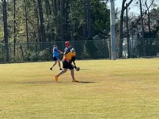 Two children playing football on a grassy field with a fence and trees in the background.