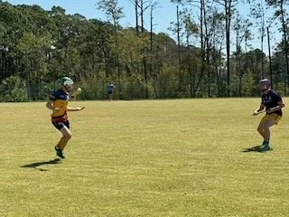 Two children playing a game of catch with a ball on a grassy field, with trees and a clear blue sky in the background.