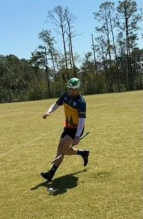 Person in athletic gear with helmet running on grassy field, trees in background.