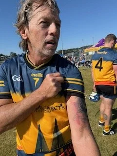 A middle-aged man with gray hair and beard wearing a blue sports jersey with the number 7 and yellow accents, holding his left arm. In the background, a person in a yellow and black sports uniform and the number 4, with a purple object (possibly a fl