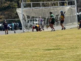 Youth baseball game with players near a net, holding a ball, and a person in a field.