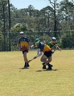 Three young boys playing lacrosse on a grassy field with trees and a fence in the background.