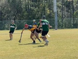 Hockey players in green and black uniforms competing for the puck on a grassy field, with a person in yellow and dark blue and a person in red helmet and shirt also involved.