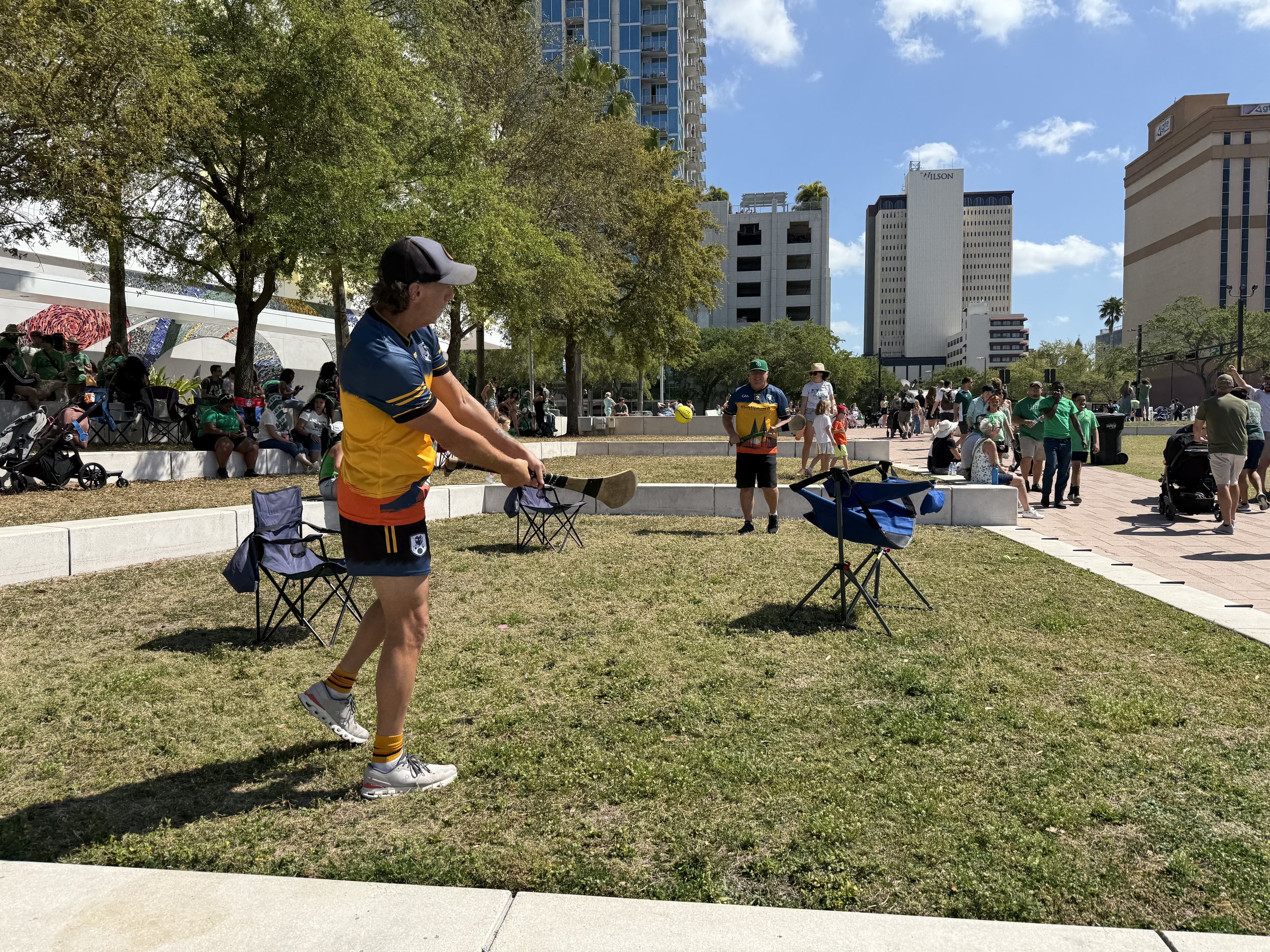 A man in a blue and yellow sports jersey and gray sneakers is playing pickleball in a park with a yellow ball. Several people are seated and standing around, some watching or socializing. There are trees, modern buildings, and a clear blue sky in the