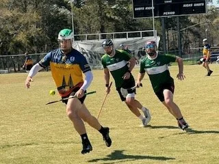 Three men playing a game of outdoor hockey on a grassy field, with a person in the background.