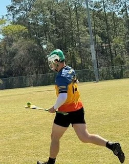 A female athlete playing lacrosse outdoors on a grassy field, holding a lacrosse stick, wearing a green helmet, colorful jersey, black shorts, and cleats.