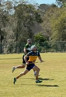 Two children playing outdoor football on a grassy field, one running ahead with a football, the other following behind.