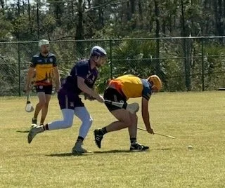 Three people playing a game on a grassy field, with one person in yellow bending over, two others nearby, one in purple and white, and one in yellow and red, all wearing helmets.