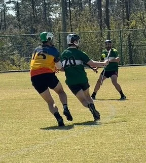 Three girls playing field hockey outdoors on a grassy field with trees in the background.