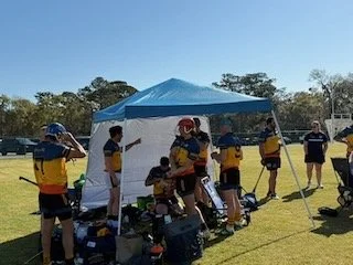 A group of children and adults in yellow and blue sports uniforms gathering under a blue and white canopy tent on a grassy field during daytime.