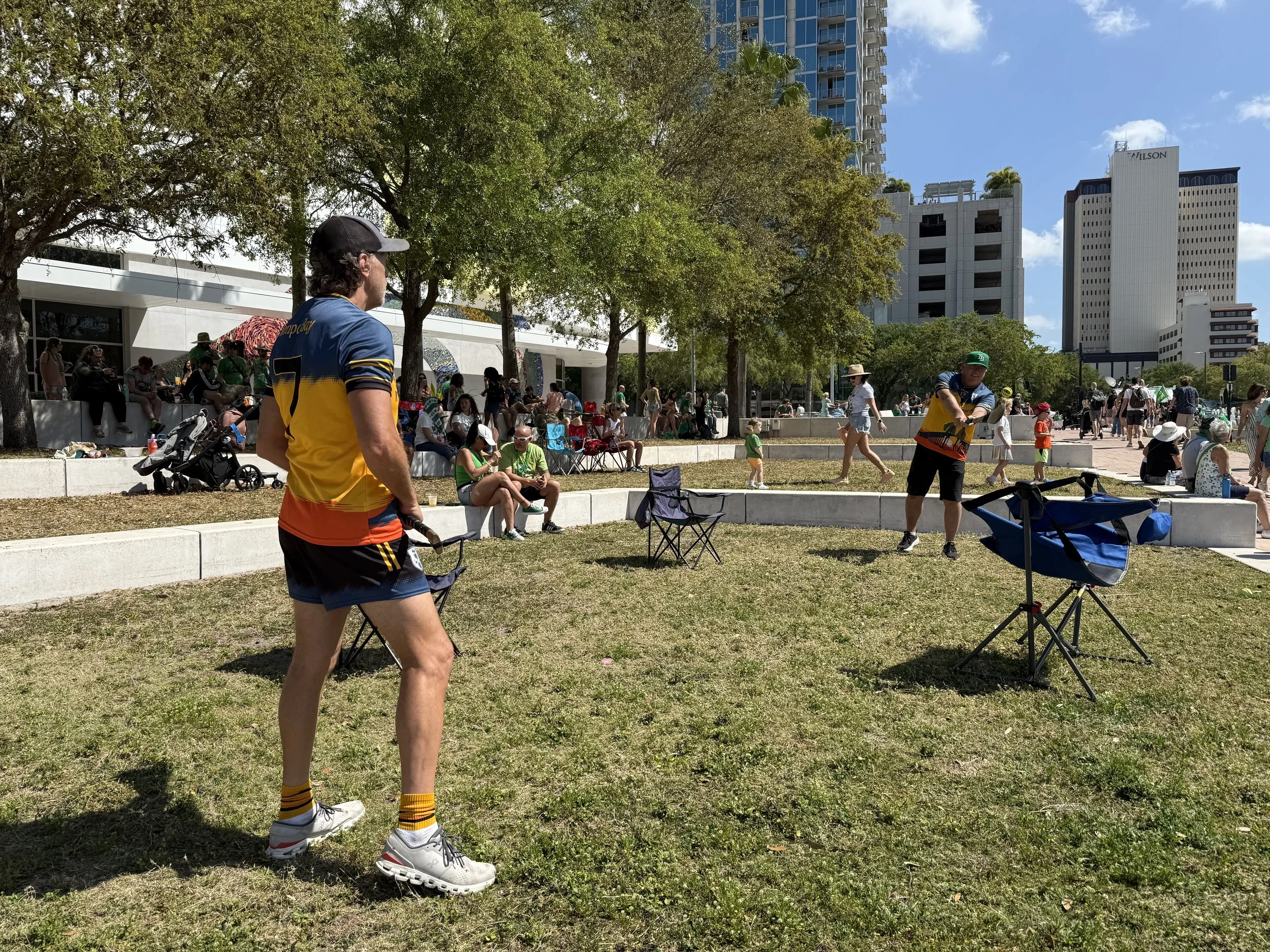 People gather in an outdoor urban park on a sunny day, some sitting on concrete benches and others walking or riding bikes, with trees, tall buildings, and a clear blue sky in the background.