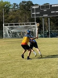 Two boys playing baseball on a grassy field, one swinging a bat and the other attempting to tag him out. In the background, there is a scoreboard and a fence.