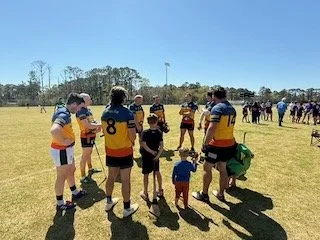 Group of hurling  players and children on a grassy field, with some players wearing yellow and blue jerseys and others in darker uniforms, under a clear blue sky.