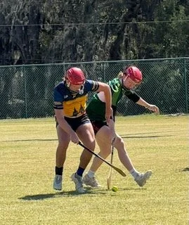 Two field hockey players in protective gear battling for possession of the ball on a grassy field.