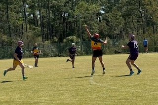 Youth flag football game on a grassy field with players in uniforms and helmets, one player jumping to catch the ball, others running or positioned around the field.