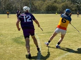 Two young hurlingl players on a grassy field during a game, with one player in a purple jersey numbered 12 and the other in a yellow and blue jersey numbered 6, running and pursuing the ball.