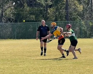 Two players in protective gear and helmets playing baseball or softball, one in yellow and the other in green, with an umpire nearby on a grassy field.