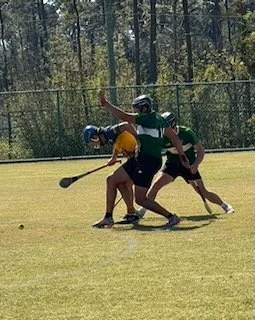 Three players in a field hockey game, two in green uniforms and one in a yellow uniform, battling for the ball on a grassy field with a chain-link fence and trees in the background.