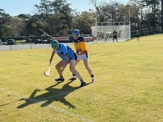 Two young players in a sports game compete for the ball on a grassy field, with a goalpost and trees in the background.