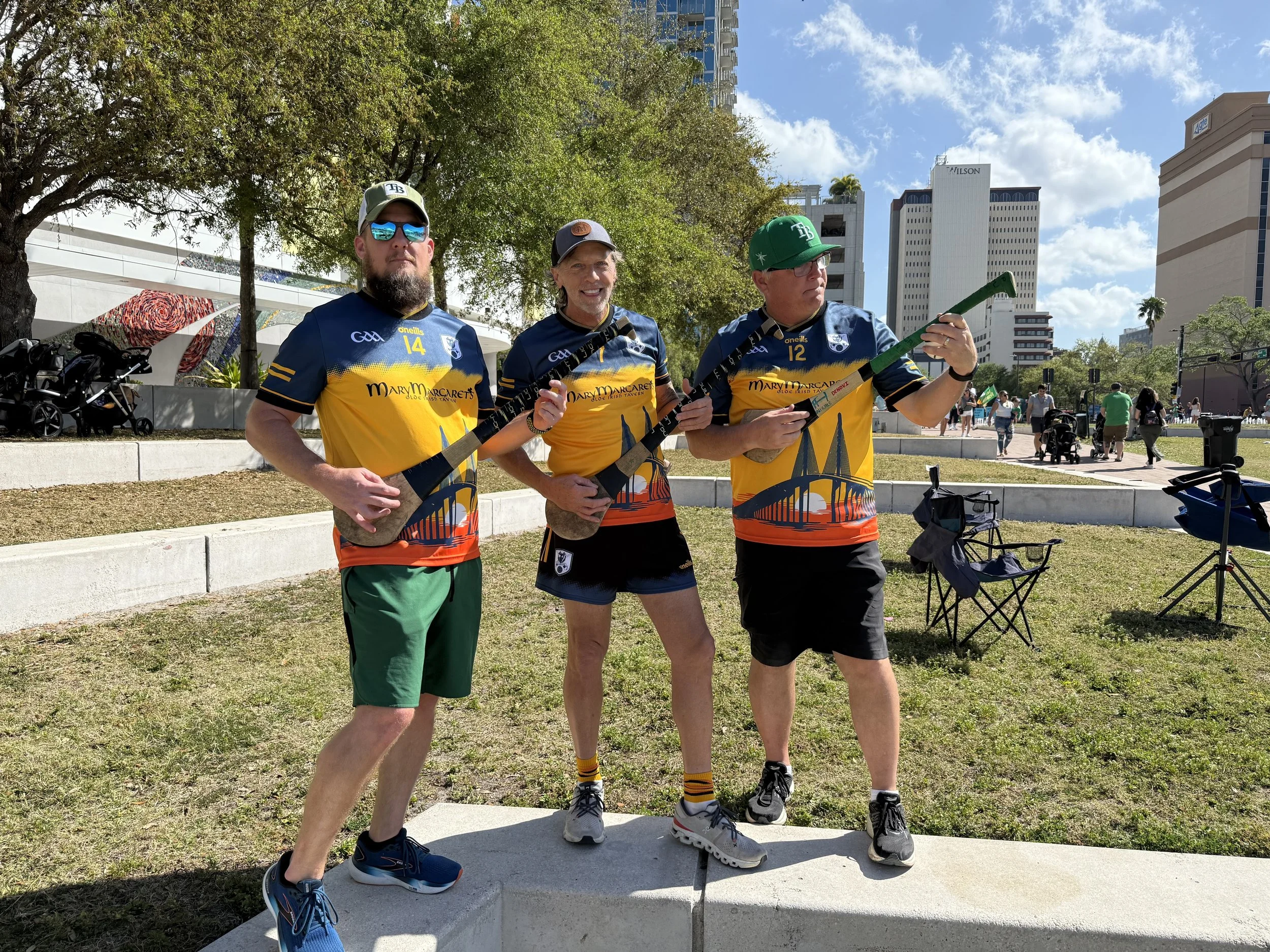 Three men standing outdoors in a city park, dressed in matching sports jerseys, holding batons, with tall buildings and trees in the background.