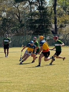 Two football players in green and yellow uniforms compete for the ball on a grassy field, with a referee and another player in the background.