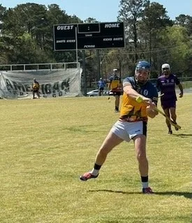 A young person playing goalie in a youth soccer game, wearing a yellow jersey, blue helmet, and white shorts, on a grassy field.