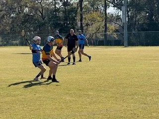 Children in sports helmets participating in a flag football game on a grassy field with an adult coach or referee.