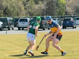 Two young children playing field hockey outdoors on a grassy field, with parked cars and trees in the background.