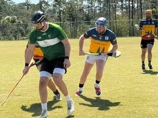 Three young lacrosse players on a grassy field during a game, wearing sports uniforms and helmets, with one player holding a lacrosse stick.