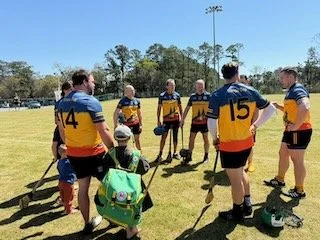 Group of young male hurling players in blue, yellow, and red jerseys standing in a huddle on a grassy field during daytime.