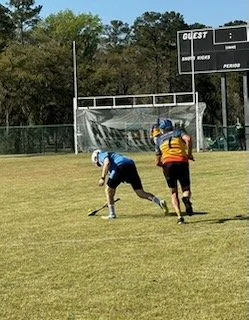 Two football players on a field during a game, one player in black and blue uniform is falling, another in red and yellow uniform is approaching.