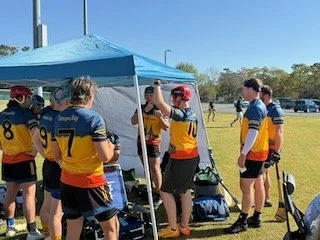 Lacrosse players gathering under a blue tent, wearing yellow and black uniforms with red helmets, on a grassy field.