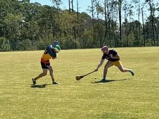 Two children playing field hockey on a grassy field with trees in the background.