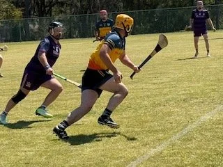 People playing a game of field hockey on a grassy field, with one person wearing a yellow helmet and an orange jersey, chasing the ball.