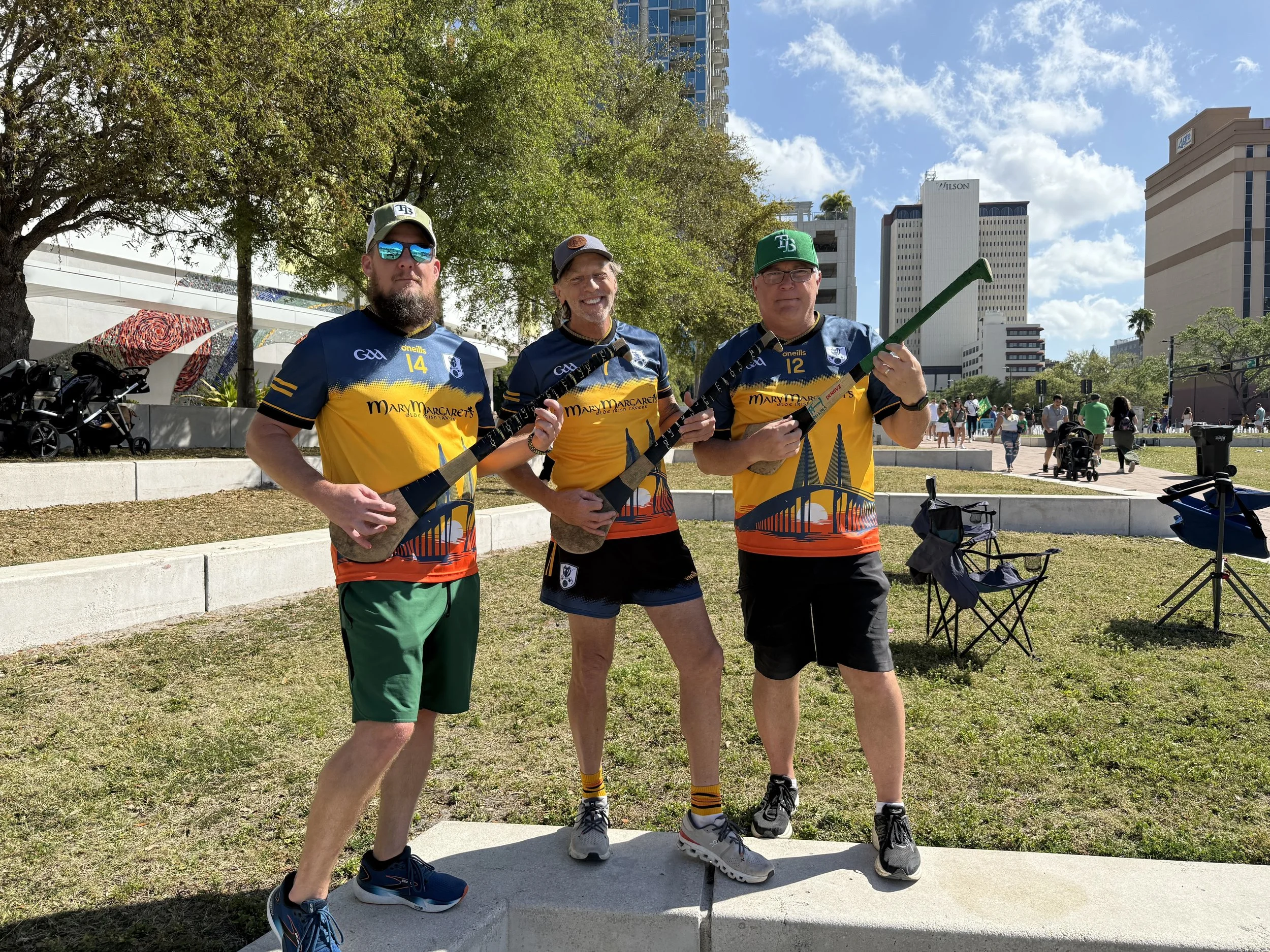 Three men standing outdoors on a sunny day, wearing blue and yellow sports jerseys, with city buildings and trees in the background. They are holding hockey sticks and posing for the photo.