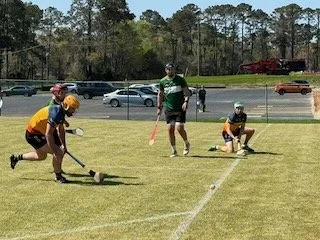 Two players in yellow and navy uniforms playing on a green field with a referee in a green shirt and shorts, biting a whistle, overseeing the game. The yellow player is sliding on the grass while the navy player is moving forward, both wearing helmet