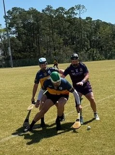 Two blindfolded individuals playing blindfolded hockey on a grassy field, with a person standing nearby and a child in the background.
