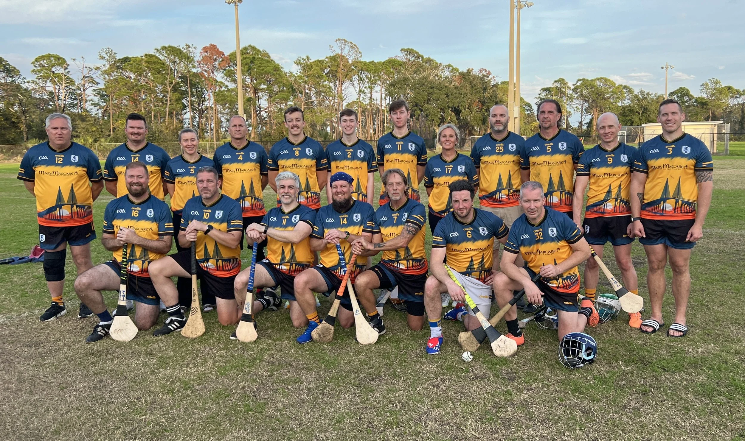 Group photo of male and female hurling players wearing blue and yellow jerseys, some holding hurleys, on a grassy field with trees and light poles in the background.