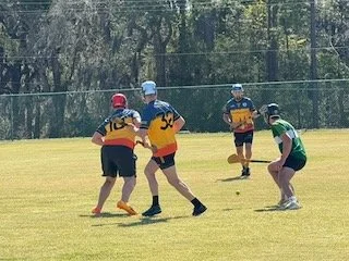 Four children playing flag football on a grassy field with trees in the background.