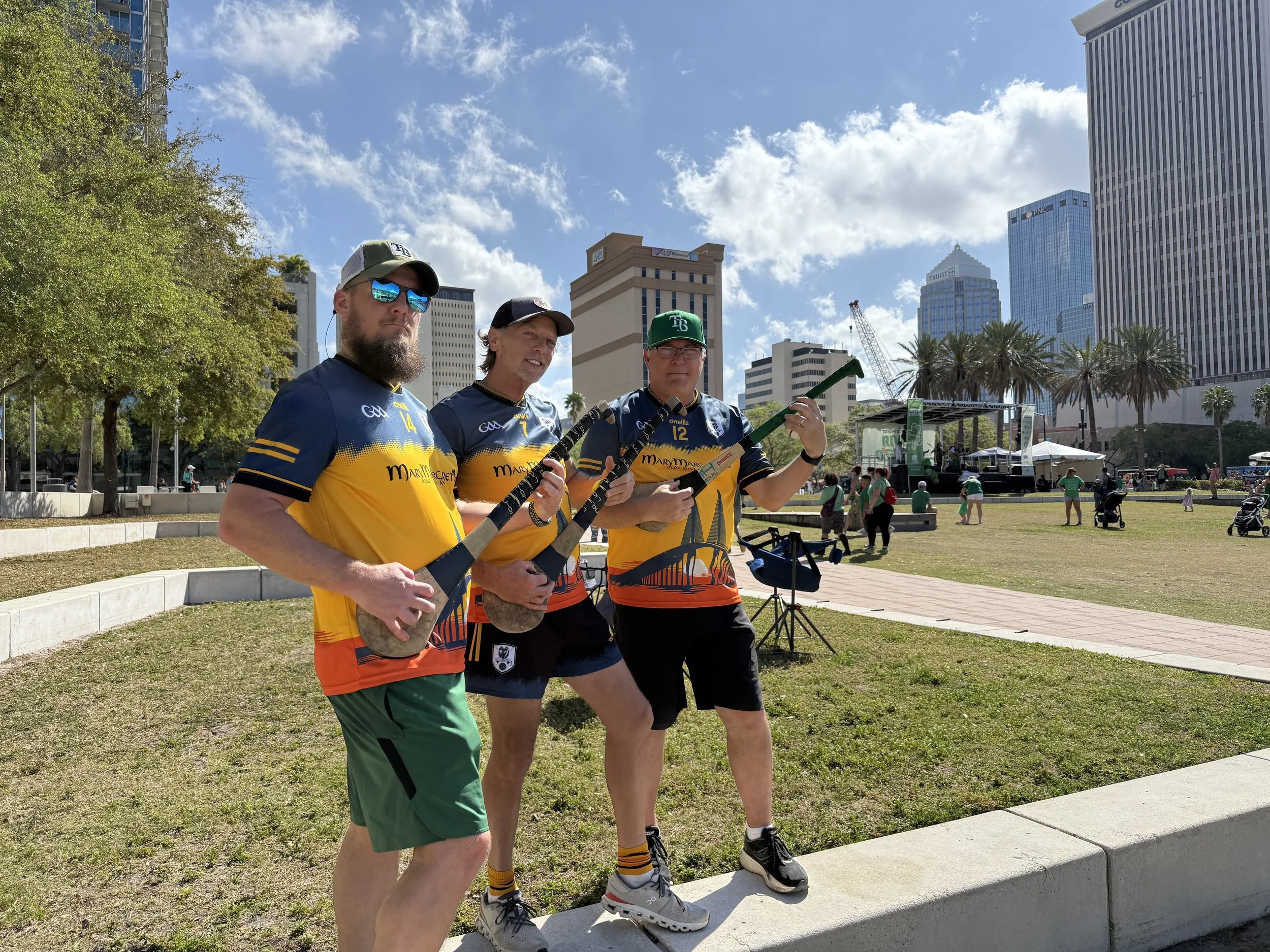 Three men hold hockey sticks while standing outdoors in a city park on a sunny day, with tall modern buildings and a stage in the background.