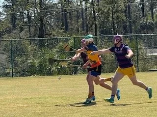 Two men playing lacrosse on a grassy field, one wearing an orange jersey and the other in purple, both running with lacrosse sticks.