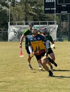 Lacrosse game with three players running on a grassy field, one wearing a blue and yellow uniform, and two in green and black uniforms, with a scoreboard and trees in the background.