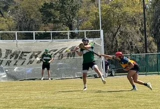 Two women playing flag football, one with a green shirt and black shorts, the other with a red helmet and yellow jersey, reaching for the flag of the woman in green on a grassy field, with a person in a green shirt and black shorts observing in the b