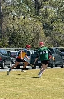 Two hurling players in helmets and jerseys compete for the ball on a grassy field with parked cars and trees in the background.