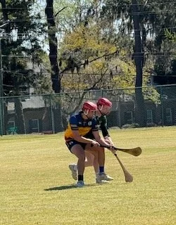 Two young boys playing hockey on a grass field with trees and a fence in the background.