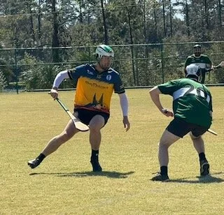 Two players in a lacrosse game on a grassy field, one in a yellow and blue jersey and the other in green and white, with a third person observing in the background.