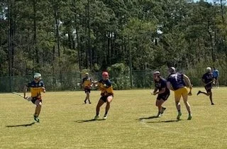 Kids playing football on a grassy field with trees in the background, wearing helmets and jerseys.