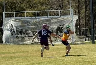 Two young boys playing football on a grassy field, with one wearing a purple jersey and the other in a yellow shirt, near a goalpost and fencing.