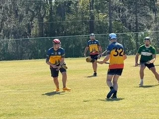 Four children playing flag football on a grassy field, wearing helmets and jerseys, with a chain-link fence and trees in the background.