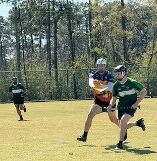 Three people playing a game of catch on a grassy field surrounded by trees, with one person wearing a helmet and two others in green jerseys.